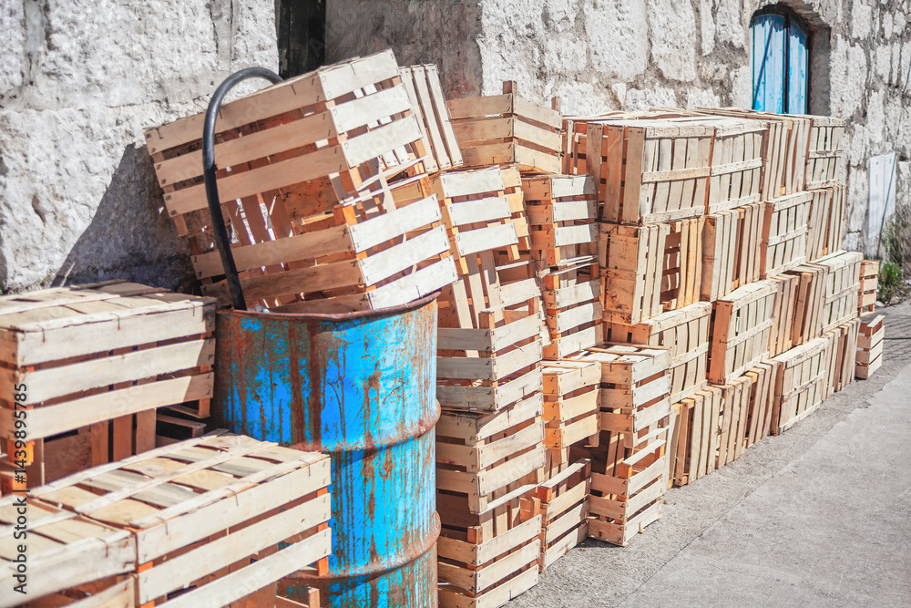 Stack of wooden crates lean against a stone wall on a concrete surface. Rusty blue barrel with a black hose sits among the crates, near a wall with a shuttered window