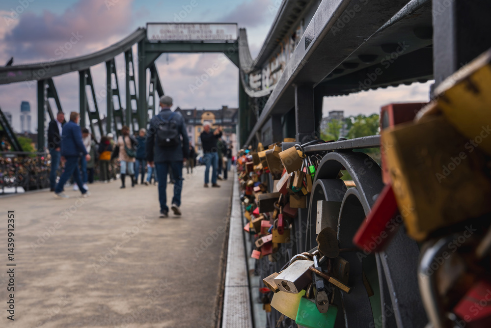 The Eiserner Steg bridge in Frankfurt, Germany, features colorful love locks on its green metal railings, with the city skyline visible under a cloudy sky.