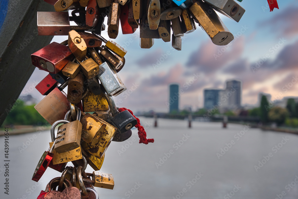 Colorful love locks adorn a metal structure, with the Main River, a bridge, and modern high rise buildings in Frankfurt, Germany, in the background.