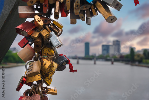 Obraz na płótnie Colorful love locks adorn a metal structure, with the Main River, a bridge, and modern high rise buildings in Frankfurt, Germany, in the background. w salonie