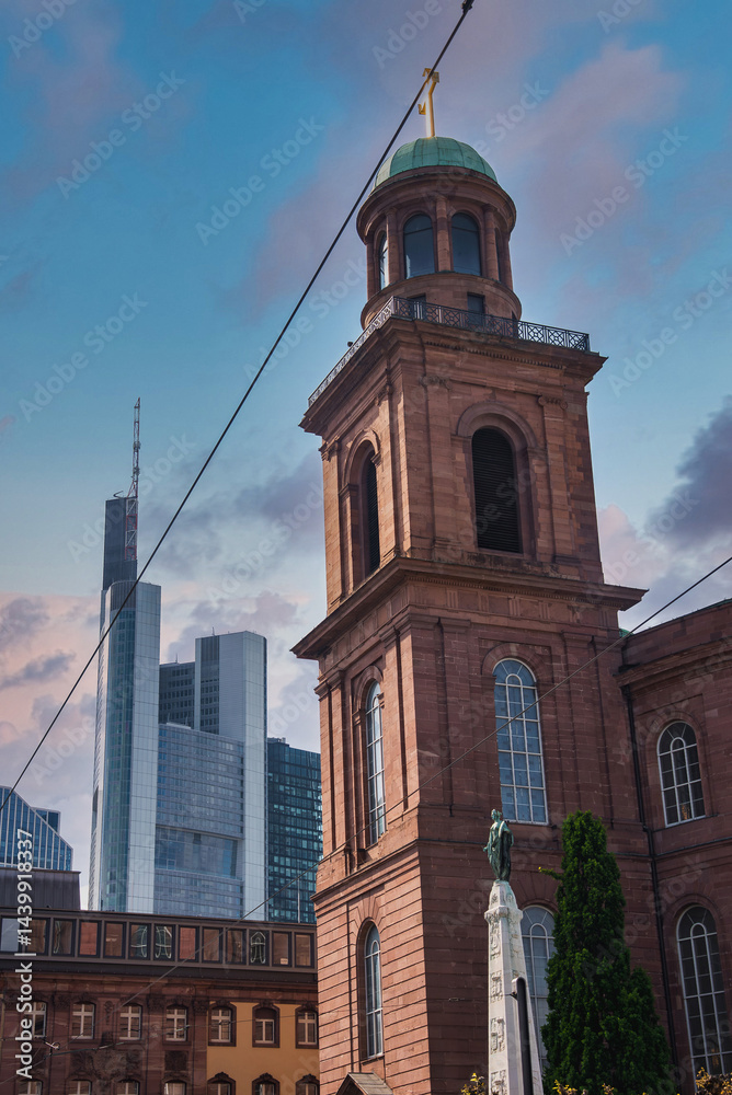 St. Paul's Church with its green dome and golden cross stands amid modern skyscrapers, including Commerzbank Tower, under a clear blue sky.