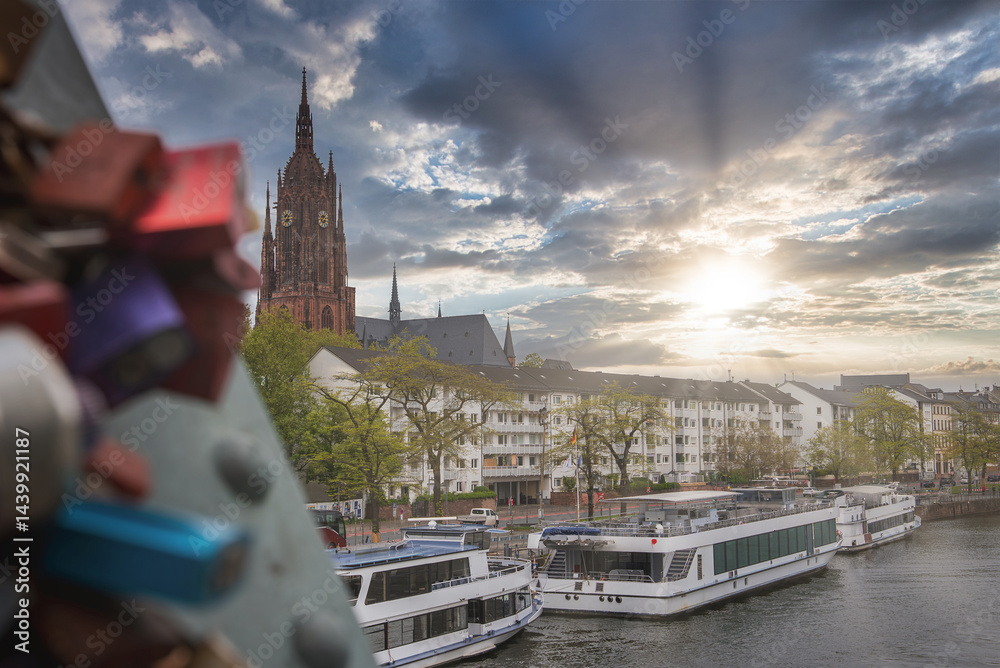 View of Frankfurt, Germany, featuring the Gothic style Frankfurt Cathedral, a bridge with love locks, the Main River, and docked riverboats.