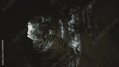 First-person view of narrow stone cave passage with visible light at the end. Mysterious underground tunnel with rough rocky walls and natural illumination.