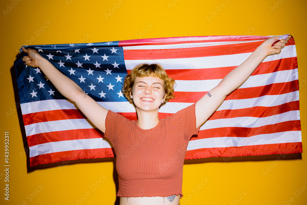 Fototapeta premium A cheerful young woman with short hair beams while holding a large American flag. The bright yellow background adds to the lively atmosphere of patriotism and joy.