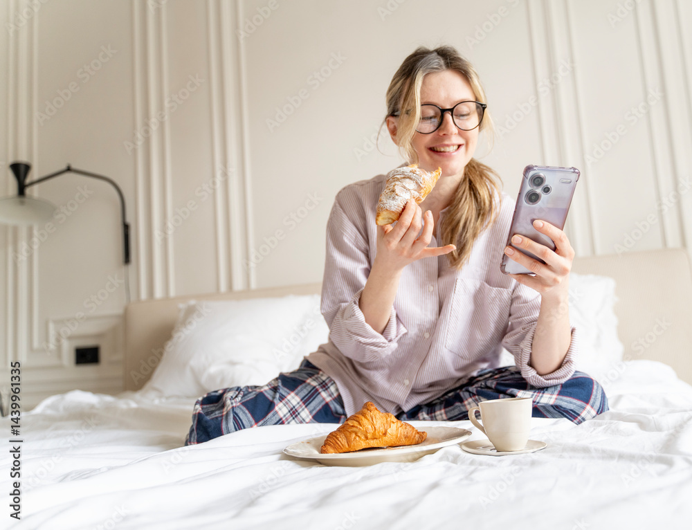 Young attractive cheerful woman in pajamas having breakfast with croissant and coffee in bed with mobile phone in hands