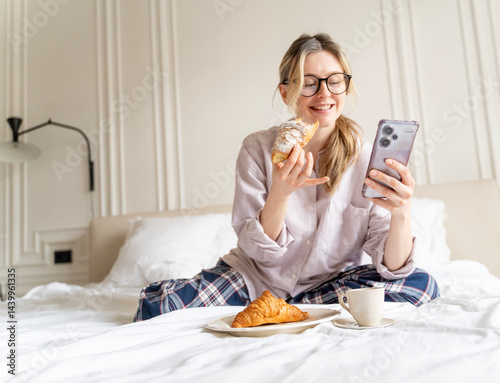 Obraz na płótnie Young attractive cheerful woman in pajamas having breakfast with croissant and coffee in bed with mobile phone in hands w salonie