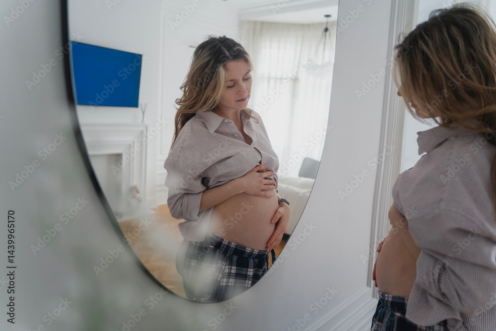 Young attractive pregnant woman looking at herself in the mirror while spending time at home