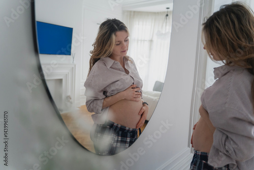 Obraz na płótnie Young attractive pregnant woman looking at herself in the mirror while spending time at home w salonie