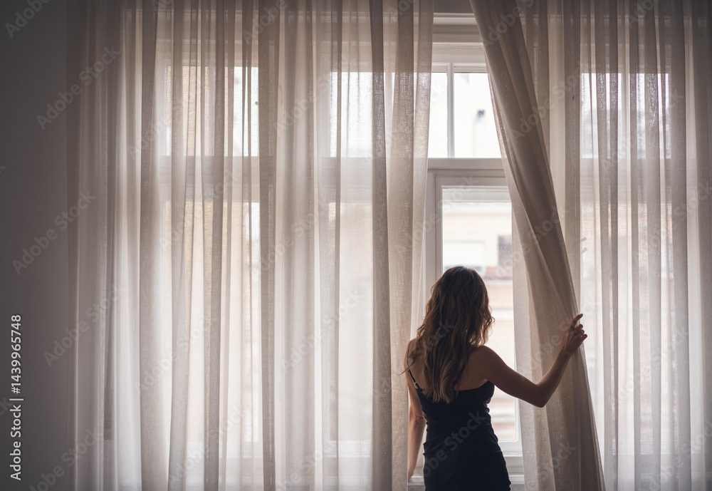 Young slim woman in black dress opening white curtains standing at window