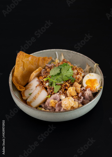 Bowl of Thai pork rice noodles soup topped with fish patties, pork crackling, boiled egg, coriander and fried garlic in black isolated background.