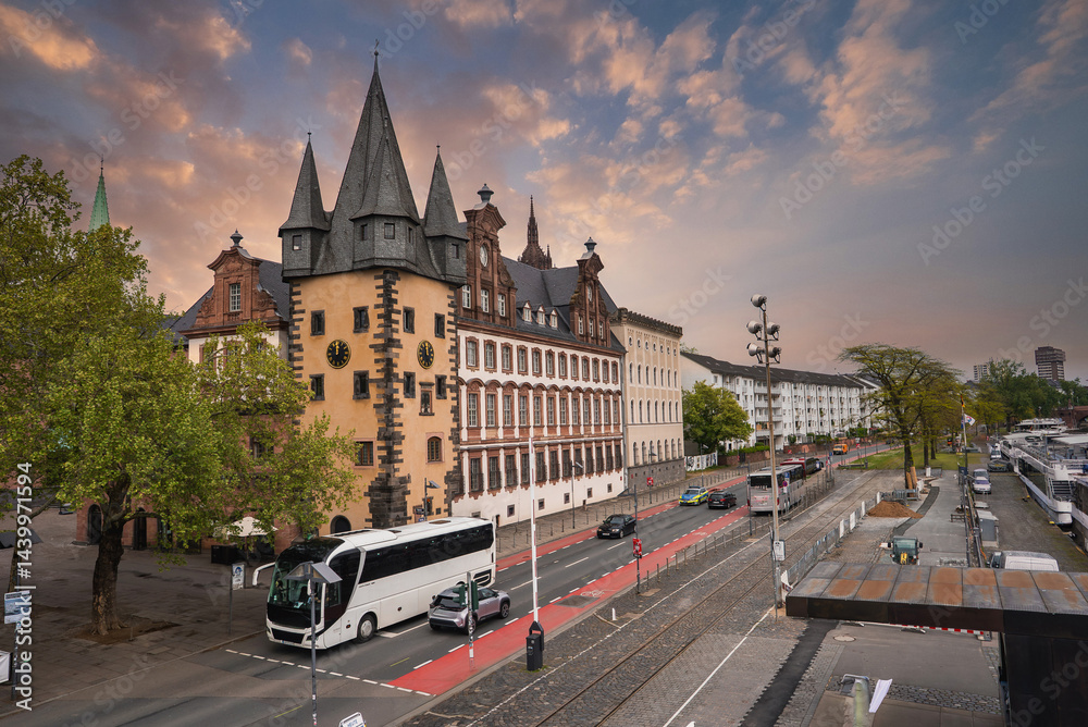 A street in Frankfurt, Germany, featuring the historic Saalhof building with pointed towers, a tree lined road, a red bike lane, and moored boats by the riverside.