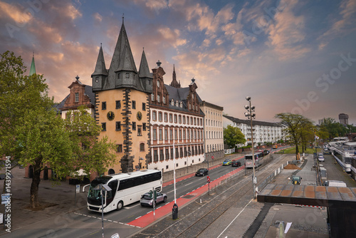 Obraz na płótnie A street in Frankfurt, Germany, featuring the historic Saalhof building with pointed towers, a tree lined road, a red bike lane, and moored boats by the riverside. w salonie