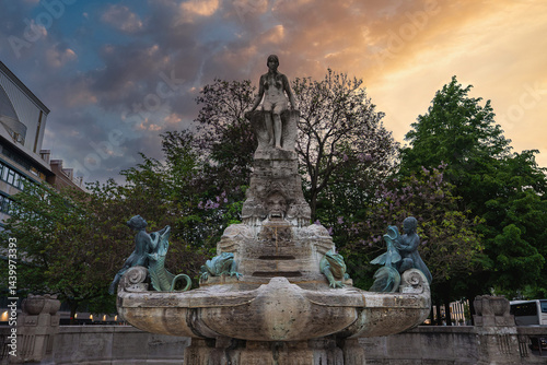 The Marchenbrunnen Fountain in Frankfurt features a central stone sculpture, bronze figures of children and animals, lush trees, and urban architecture. photo
