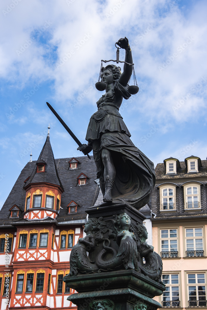 Obraz premium The Fountain of Justice statue in Frankfurt's Romerberg square with Lady Justice, surrounded by half timbered buildings under a bright blue sky.