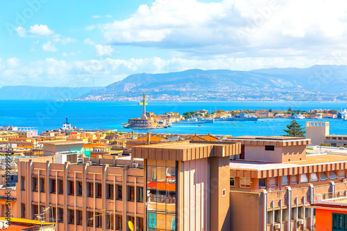 View of Messina, Sicily, with modern buildings in the foreground, the iconic Madonna della Lettera statue, and the Strait of Messina in the background under a bright, partly cloudy sky