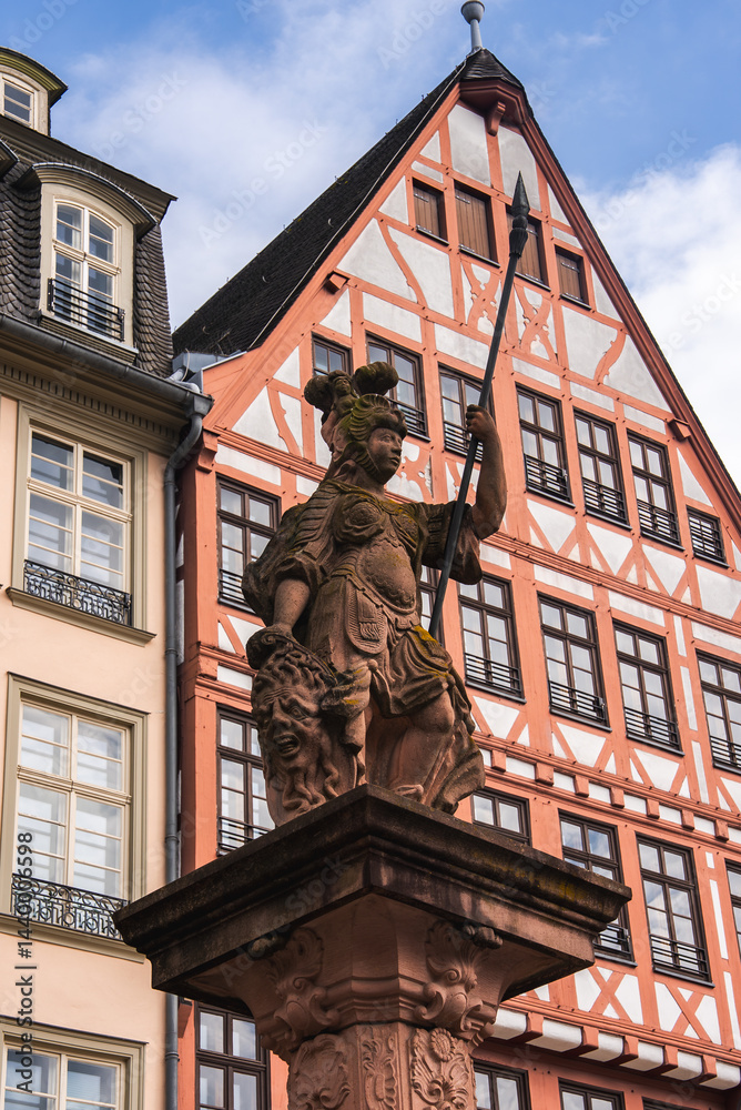 Obraz premium A detailed statue of a warrior holding a spear stands on an ornate pedestal, with a red and white half timbered building and modern architecture in Frankfurt.
