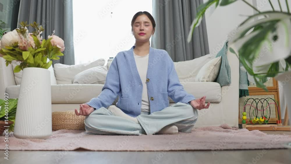 Asian young woman sits cross-legged on a carpet, meditating with eyes closed in a peaceful living room environment. Mindfulness, inner calm, and a wellness lifestyle, surrounded by plants