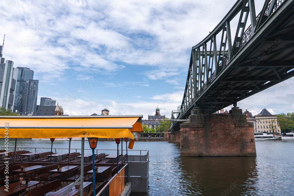 The Eiserner Steg bridge spans the Main River in Frankfurt, Germany, with steel and stone details, a docked yellow roofed boat, and city skyscrapers.