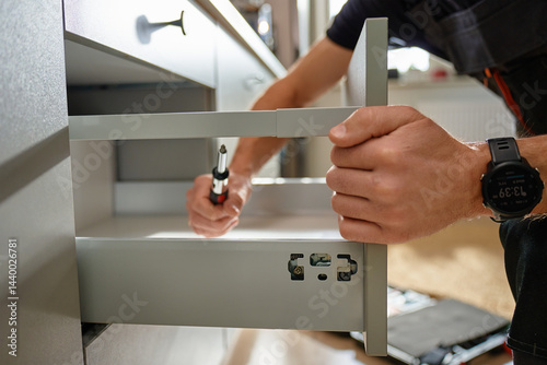 Obraz na płótnie Man assembling kitchen drawer in modern cabinet using screwdriver. Handyman installing and adjusting drawer slide mechanism in contemporary kitchen. Furniture installation w salonie