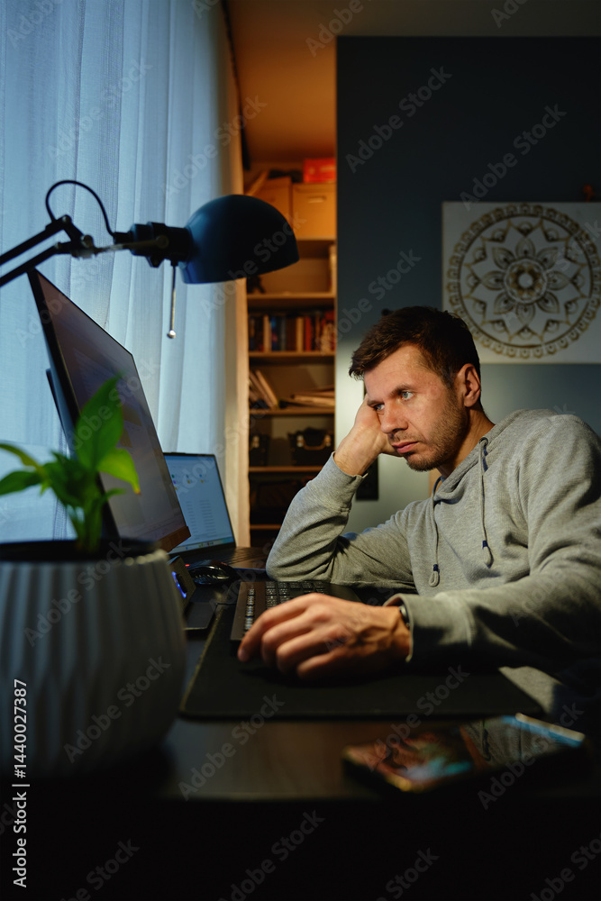 Unfocused man sitting at desk with hand on face, looking at screen. Freelancer distracted at home office. Concept of procrastination, overwork and digital burnout