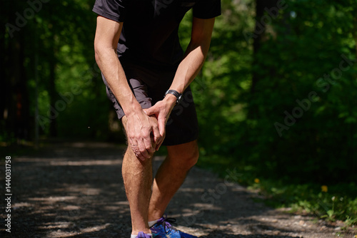 Fototapeta Naklejka Na Ścianę i Meble -  Runner holding knee in pain during workout in summer park. Man has injury while running. Male athlete suffering from knee pain after sport exercises