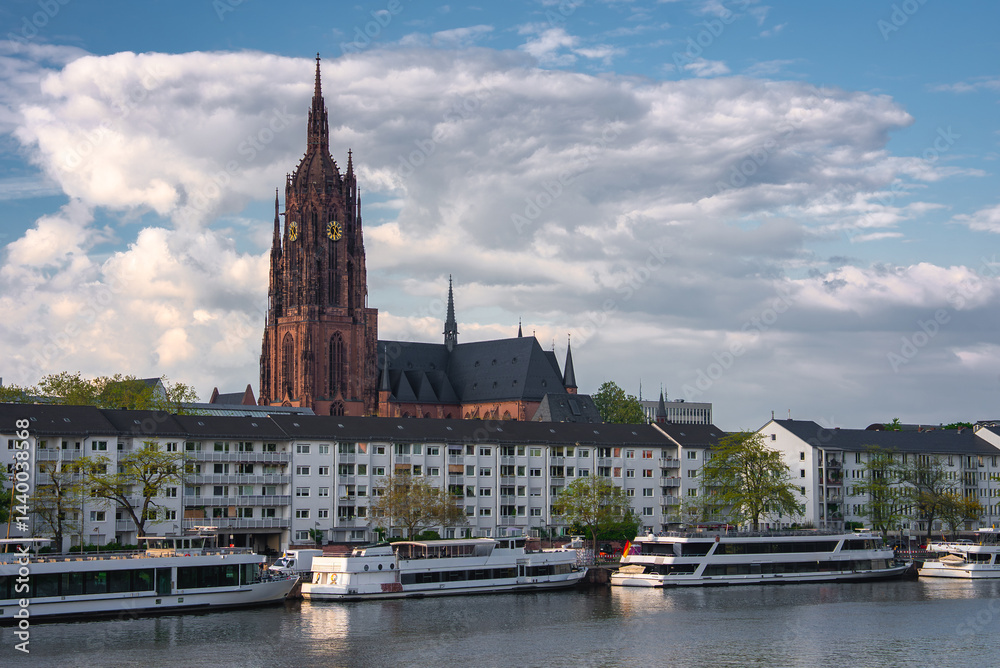 The Frankfurt Cathedral's Gothic spire rises against a cloudy sky, with the Main River, docked boats, and modern residential buildings in the foreground.