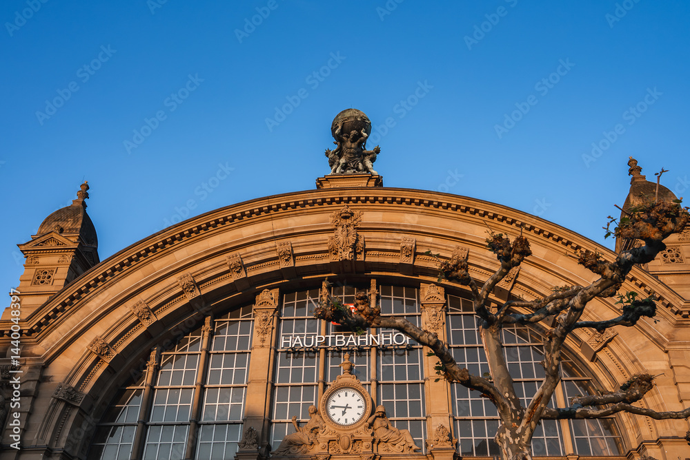 The facade of Frankfurt Hauptbahnhof in Germany features a grand arched window, a central clock, Atlas statue, smaller sculptures, and a clear blue sky.
