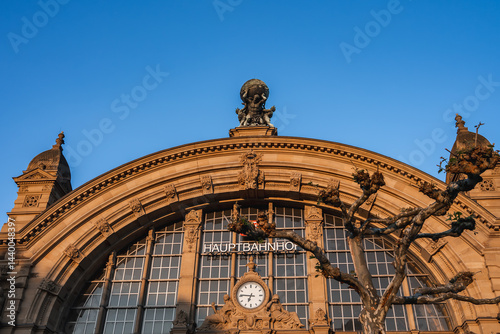 The facade of Frankfurt Hauptbahnhof in Germany features a grand arched window, a central clock, Atlas statue, smaller sculptures, and a clear blue sky.