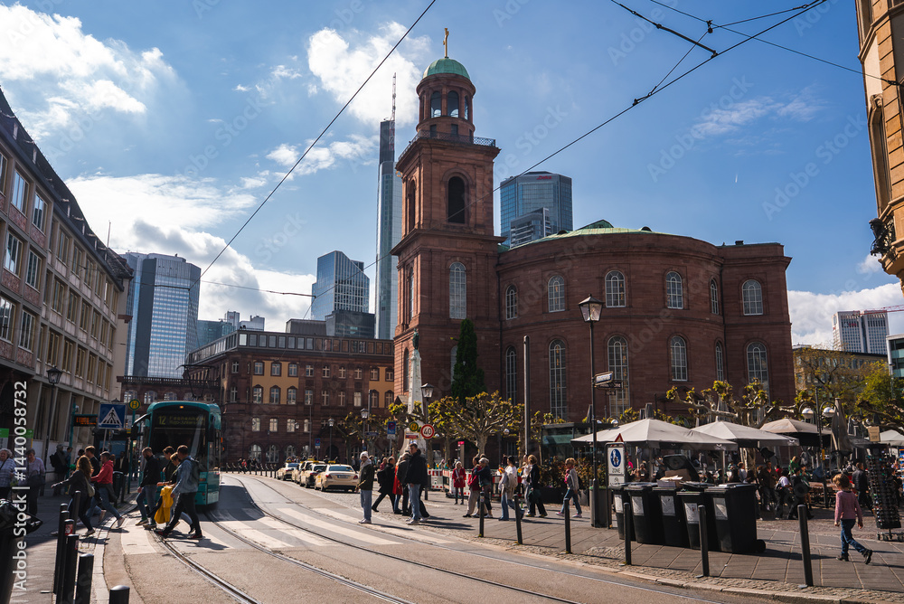 Bustling street scene in Frankfurt with St. Paul's Church, modern skyscrapers, pedestrians, a tram, outdoor seating, and clear blue skies.