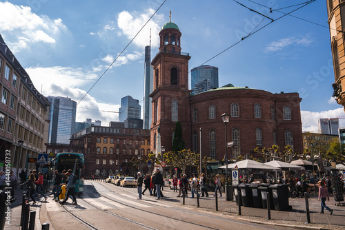 Obraz na płótnie Bustling street scene in Frankfurt with St. Paul's Church, modern skyscrapers, pedestrians, a tram, outdoor seating, and clear blue skies. w salonie