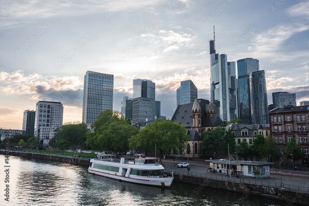 Skyline of Frankfurt, Germany, featuring modern skyscrapers like Main Tower, a historic church, the River Main, a docked boat, and green trees.
