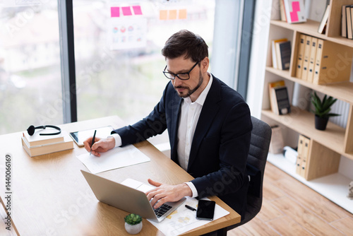 Young adult male in formal attire working in office, using laptop, taking notes on paper, conducting business task. Office environment has natural light, bookshelves, and work materials.