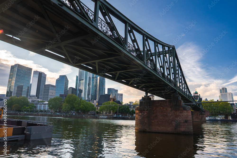 The Eiserner Steg bridge spans the Main River in Frankfurt, Germany, with intricate ironwork, stone piers, and a modern skyline in the background.