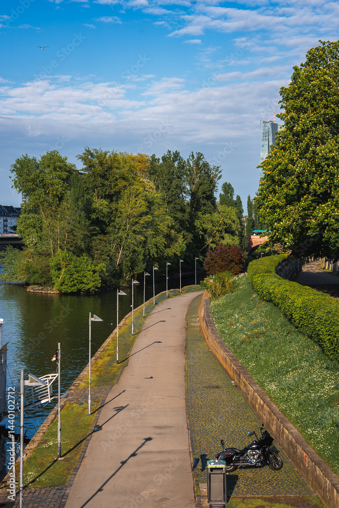 A paved riverside pathway in Frankfurt, Germany, bordered by greenery and a calm river. A motorcycle is parked near the path, with a skyscraper visible.