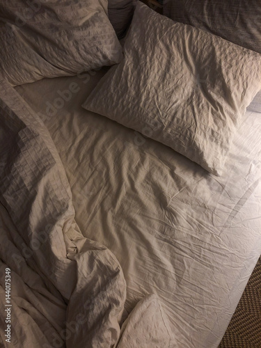 Cozy bedroom night setting featuring white wrinkled white linen and fluffy pillows on a wooden bed, capturing a warm and inviting restful atmosphere. Inviting view of fresh and clean bedding.