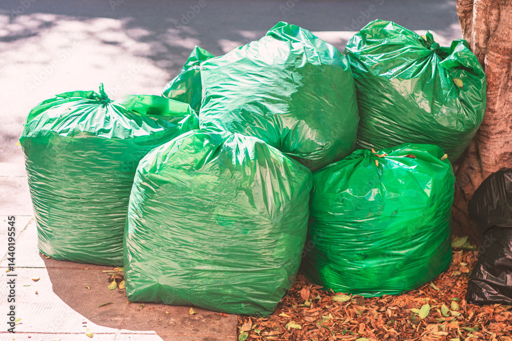Overflowing green garbage bags are situated on a paved surface, amongst fallen leaves. Full refuse bags, tied rest on the ground