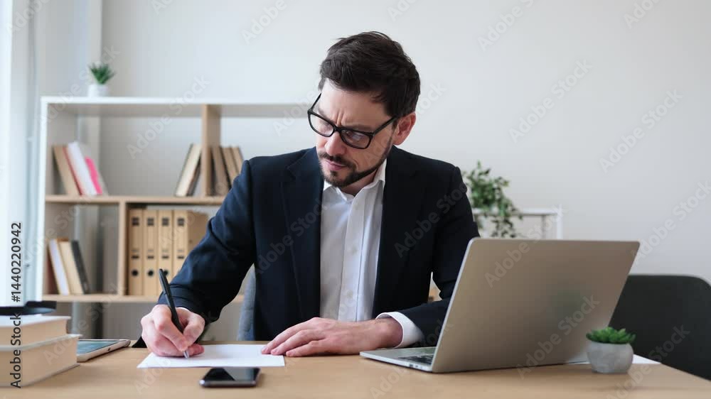 Businessman in mid-30s thinking and writing notes in office with laptop and books. Businessman in mid-30s thinking and writing notes in office with laptop and books.