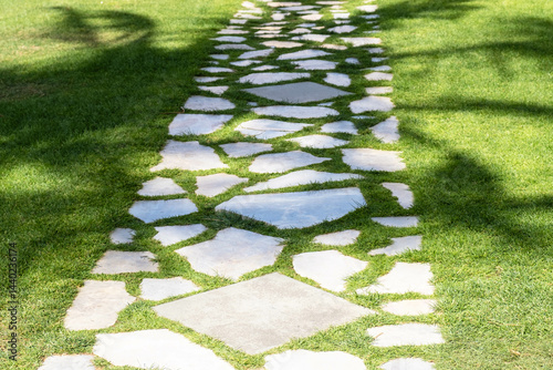 Stone pathway meandering through a lush green lawn, creating a natural walkway. Sunlight casts shadows of nearby trees, enhancing the peaceful outdoor garden setting