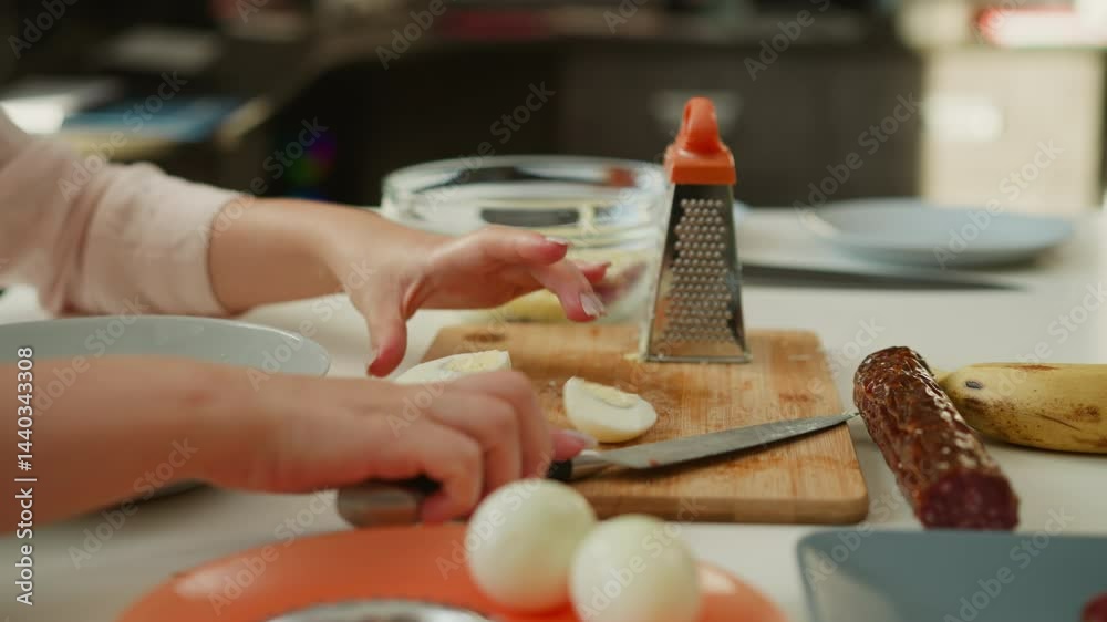 Hand of cook slicing boiled egg in half on wooden cutting board with sharp knife, grater and glass bowl nearby on kitchen table during meal preparation in bright home environment