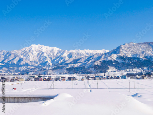 真っ白な雪に覆われた山と、その麓に広がる小さな集落を捉えた冬の風景。澄んだ青空が美しいコントラストを演出しています。