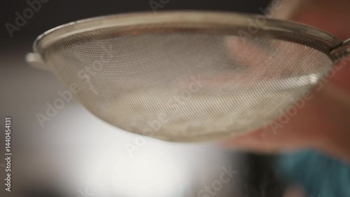 close up hand tapping sieve to sift flour into glass bowl in kitchen setting with soft blurred background showing child silhouette, mixing whisk and rolling pin scattered on counter top