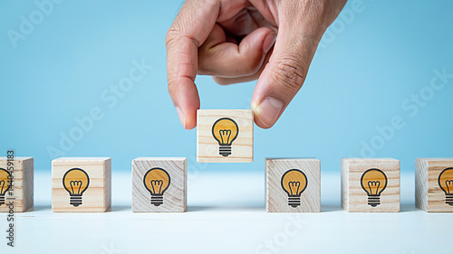 Hand Placing Wooden Cube with Yellow Light Bulb Icon on White Surface, Surrounded by Identical Beige Blocks with Black Icons