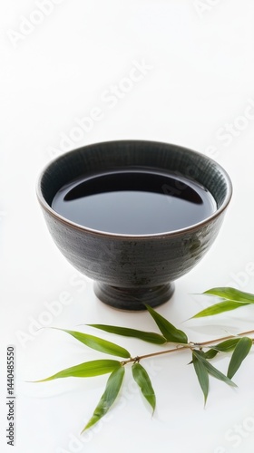 Dark ceramic bowl filled with black liquid accompanied by green bamboo leaves on white