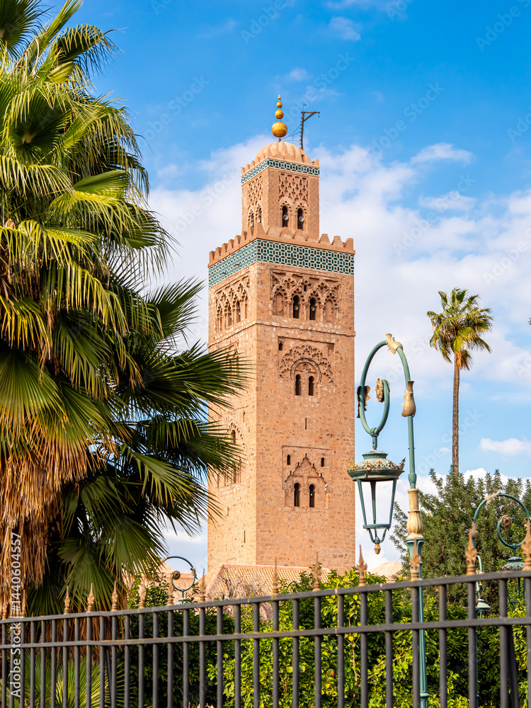Fototapeta premium Koutoubia Mosque minaret in Marrakesh, Morocco, surrounded by palm trees under bright blue sky, showcasing historic Islamic architecture, cultural heritage, religious significance and vibrant scenery.