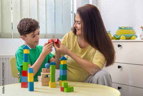 Happy young boy and woman playing with colorful wooden blocks at home. Concept of early childhood education, autism therapy, ADHD support, child psychology, occupational therapy, family connection. 