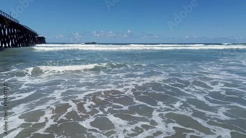 Drone shot, zooming in and out over gentle waves rolling onto the Rosarito Beach pier on a beautiful day.