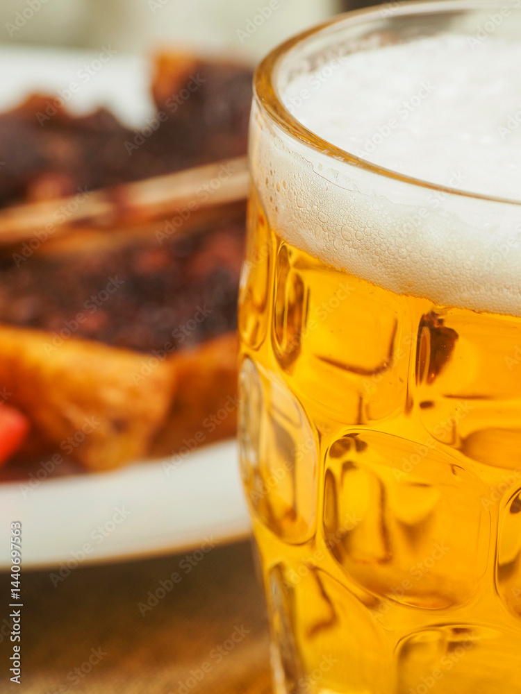 Glass of lager beer in focus, cooked beef T bone steak meal out of focus. Premium high quality meat served on a plate with wedges and fresh tomato. Fine expensive well done meal.