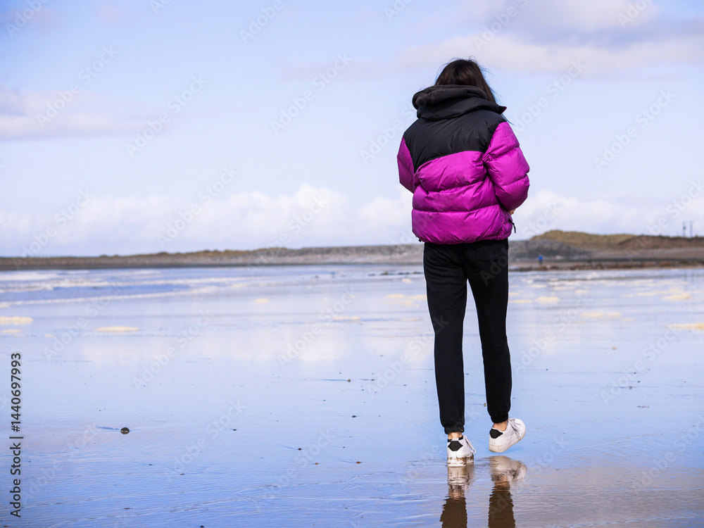 Teenager girl in purple jacket walking on a wet sandy beach which reflects blue cloudy sky. Travel to seaside concept. Relaxing activity by the ocean. Selective focus.