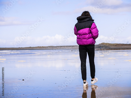 Obraz na płótnie Teenager girl in purple jacket walking on a wet sandy beach which reflects blue cloudy sky. Travel to seaside concept. Relaxing activity by the ocean. Selective focus. w salonie