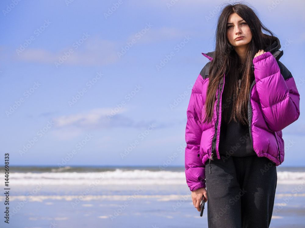 Portrait of a young teenager girl on a ocean beach. Model with long dark hair in purple jacket holding a smart phone, blue sky and water background.. Selective focus. Travel and tourism concept.